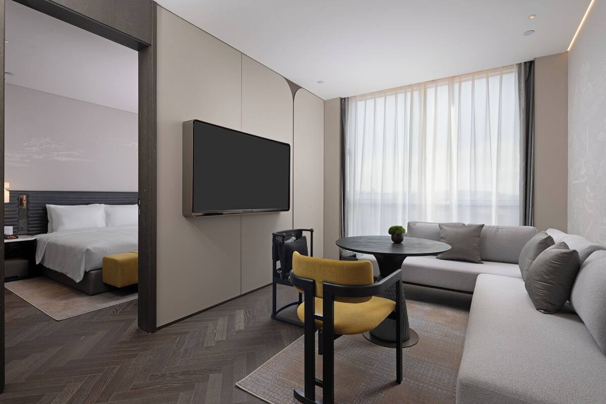 A modern hotel room with a bed, a sofa, a TV, and a dining area.
