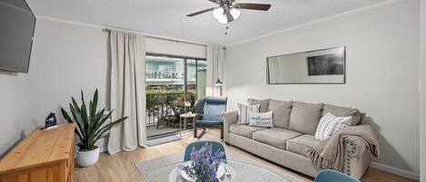 A modern living room with a sofa, a coffee table with a vase of flowers, a ceiling fan, and a view of the outside balcony.