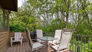 A wooden deck with a glass table and four chairs, surrounded by a metal railing and lush greenery.