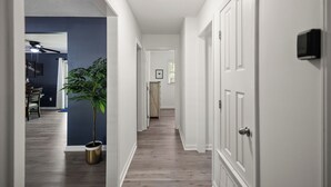 A hallway with wooden flooring, white walls, and a ceiling light. There is a potted plant and a door with a security camera. - NEW - Moody BBQ Retreat w/Covered Patio (Jackson)
