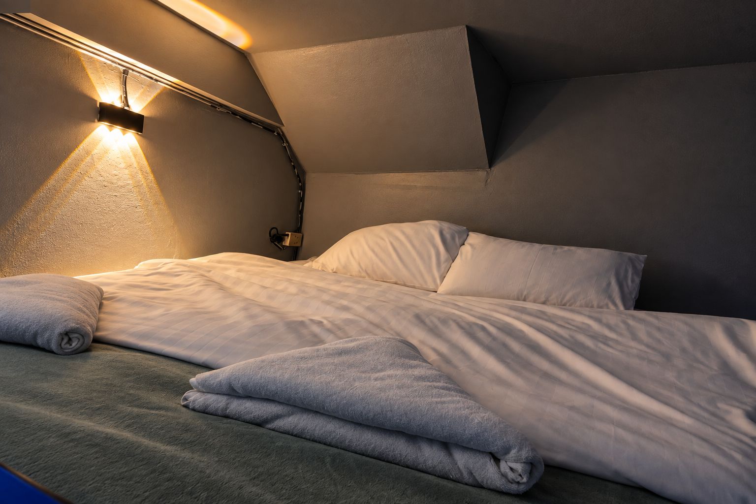 Un lit bien fait, avec des oreillers et une couverture pliée, un luminaire fixé au mur et un mur sobre.