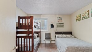 A compact bedroom with bunk beds, a single bed, a chair, a wall-mounted air conditioner, and a small window with blinds. - Beach Block steps to sand and many more attractions making this the perfect spot (Wildwood)