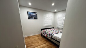 Une chambre avec un plancher en bois, un lit recouvert d’une couverture colorée, des murs blancs et un tableau accroché au mur.