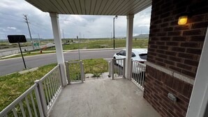 Une véranda couverte avec un mur en briques, une balustrade blanche et une vue sur une route où circulent des voitures.