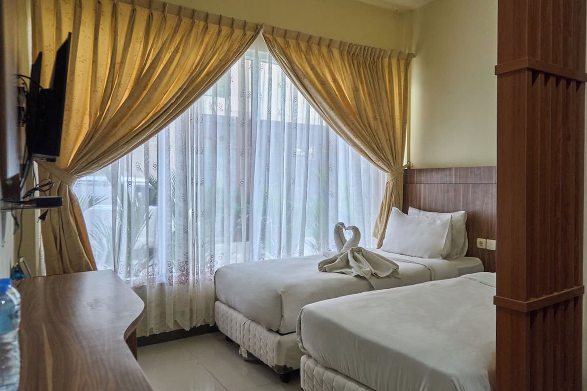 A hotel room with two beds, a wooden headboard, a TV mounted on the wall, and a window with sheer curtains.