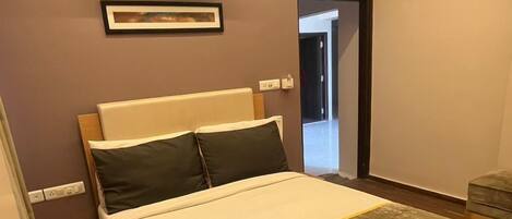 A modern hotel room with a large bed, a ceiling fan, an air conditioning unit, a framed picture on the wall, and a wooden floor.