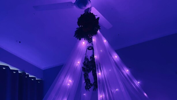Habitación con una cama bajo un dosel de luces moradas, un ventilador de techo y un cabecero oscuro.
