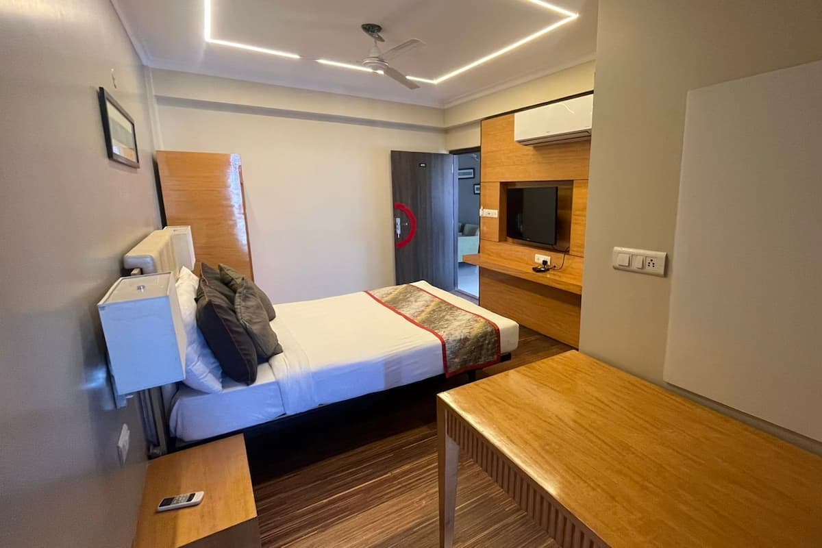 Quarto de hotel compacto com uma cama de solteiro, uma escrivaninha de madeira, uma televisão e um ventilador de teto.
