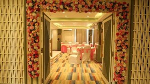 Un pasillo de hotel con un arco floral, sillas blancas con cintas rosas y una alfombra de diseño colorido.