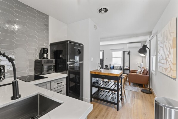 Eine moderne Küche mit Einbauofen, Kühlschrank und Kaffeemaschine. Im Wohnbereich befindet sich ein Esstisch mit Stühlen und ein Sofa.