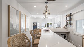 A modern dining area with a long white table, wicker chairs, and a central island with a bar.