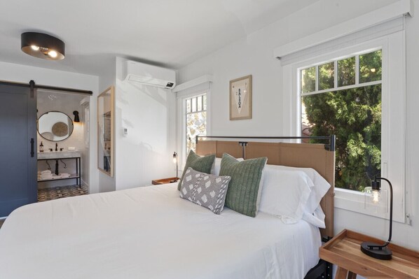 Un dormitorio con una cama grande, una mesita de noche con lámpara, una ventana con vistas a árboles y un baño con espejo y lavamanos.