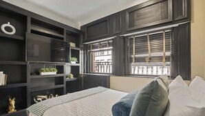 A bedroom with a large bed, dark wood shelving, a window with blinds, and a small plant on the shelf.