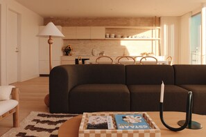 Uma sala de estar com um sofá marrom escuro, uma mesa de centro de madeira com livros, uma luminária e, ao fundo, uma cozinha com parede de tijolos e armários de madeira.