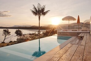 An infinity pool with a view of the sunset over a beach and mountains.