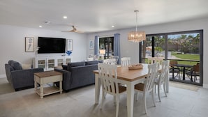 A modern living room with a sofa, TV, dining table, and chairs, with a large window and ceiling fan. - Blind Pass C106  | Upgraded Ground Floor Residence at Blind Pass | Steps to Pool (Sanibel)