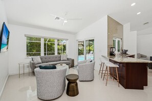 A modern living space with a large window, a grey sofa, a coffee table, a bar area, and a kitchen.