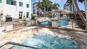 Zona de hidromasaje con piscina y sillones de descanso, rodeada de palmeras y un edificio blanco.