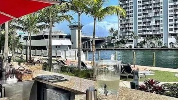 Un bar frente al agua con vistas a un puerto deportivo, palmeras y un rascacielos al fondo.