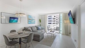 Una sala de estar moderna con un sofá gris, una mesa de comedor con sillas, una araña y un ventanal con vistas a edificios.