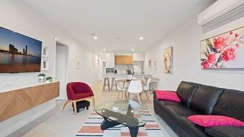 A modern living room with a glass coffee table, a leather sofa with red cushions, a dining area with white chairs, and a kitchen in the background.