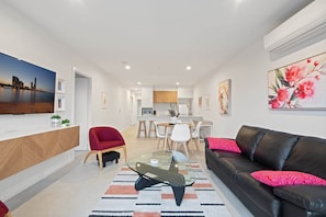 Un salon moderne avec une table basse en verre, un canapé en cuir avec des coussins rouges, un coin repas avec des chaises blanches et une cuisine en arrière-plan.