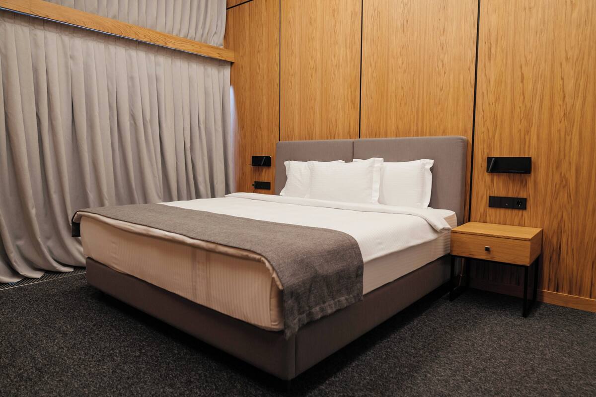 Una cama bien hecha con cabecera gris, una mesita de noche de madera con cajón, y un panel de pared con un interruptor.