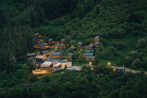 A cluster of log cabins surrounded by dense forest, with a central building featuring a large porch and a circular light fixture. - Qazaq Auyl (Almaty)