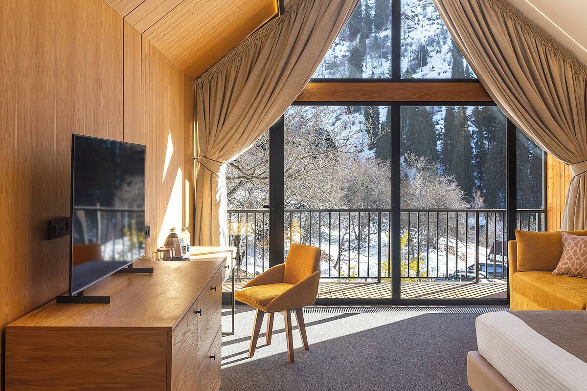 Un dormitorio estilo loft moderno con un ventanal grande, televisor de pantalla plana, muebles de madera y un balcón con vistas a árboles.