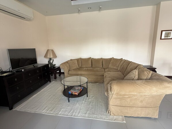 A living room with a sectional sofa, a glass coffee table, a TV, a lamp, and a dark wooden dresser.
