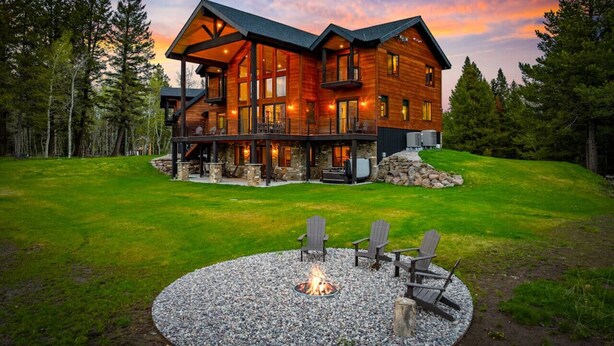 A large, multi-story log cabin with a stone foundation, a fire pit, and outdoor seating.