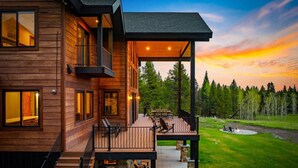 A modern wooden house with a deck, outdoor seating, and a view of a forested area.