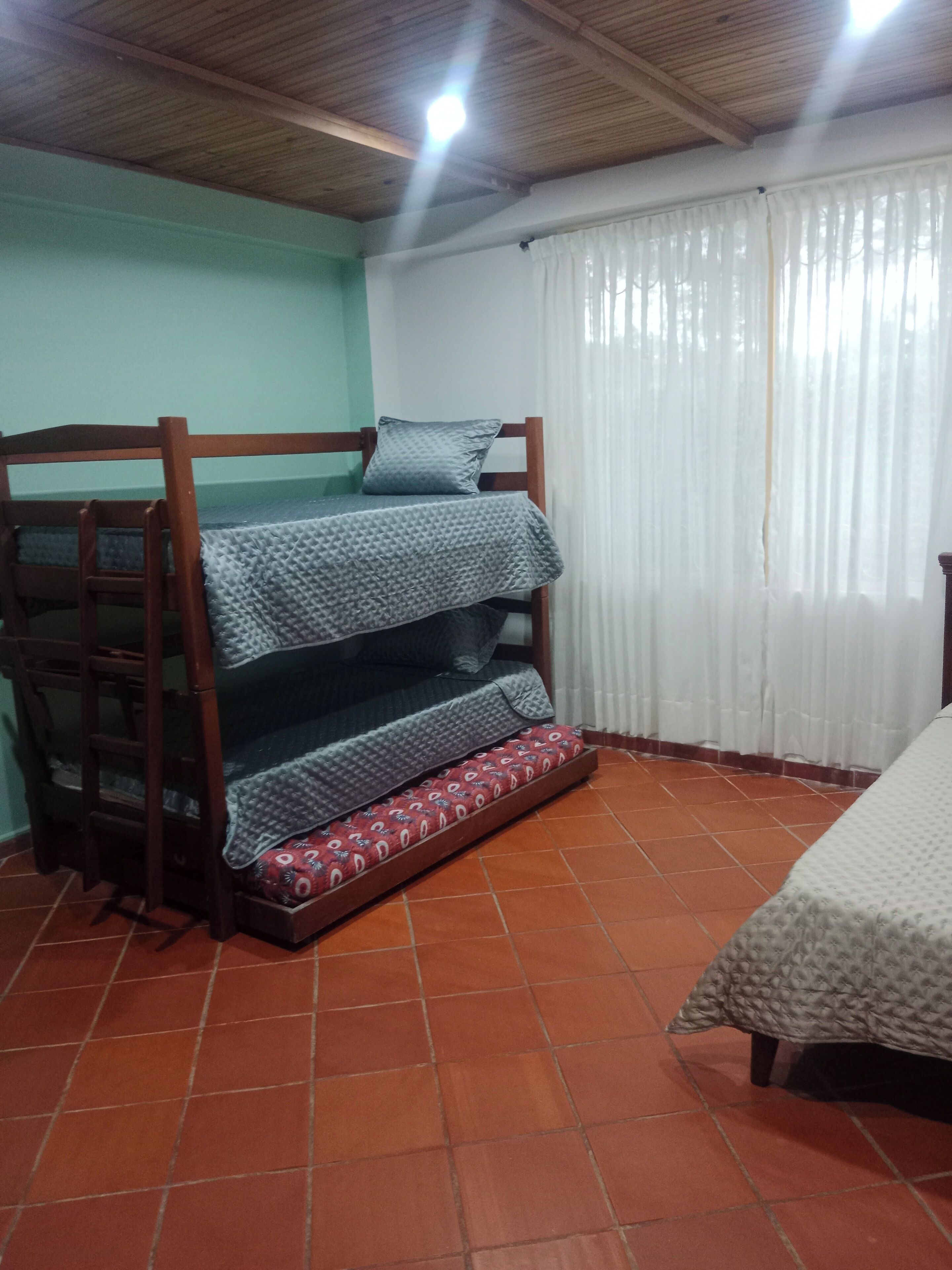 Un lit superposé avec un matelas à motifs, une échelle en bois et un oreiller. La pièce a un sol carrelé et une fenêtre avec des rideaux.