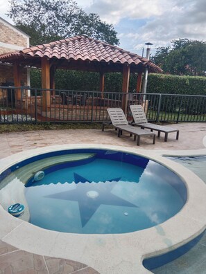 Ein sternförmiger Pool, umgeben von einer gefliesten Liegefläche. Zwei Liegestühle und ein Gazebo mit einem gefliesten Dach sind vorhanden.