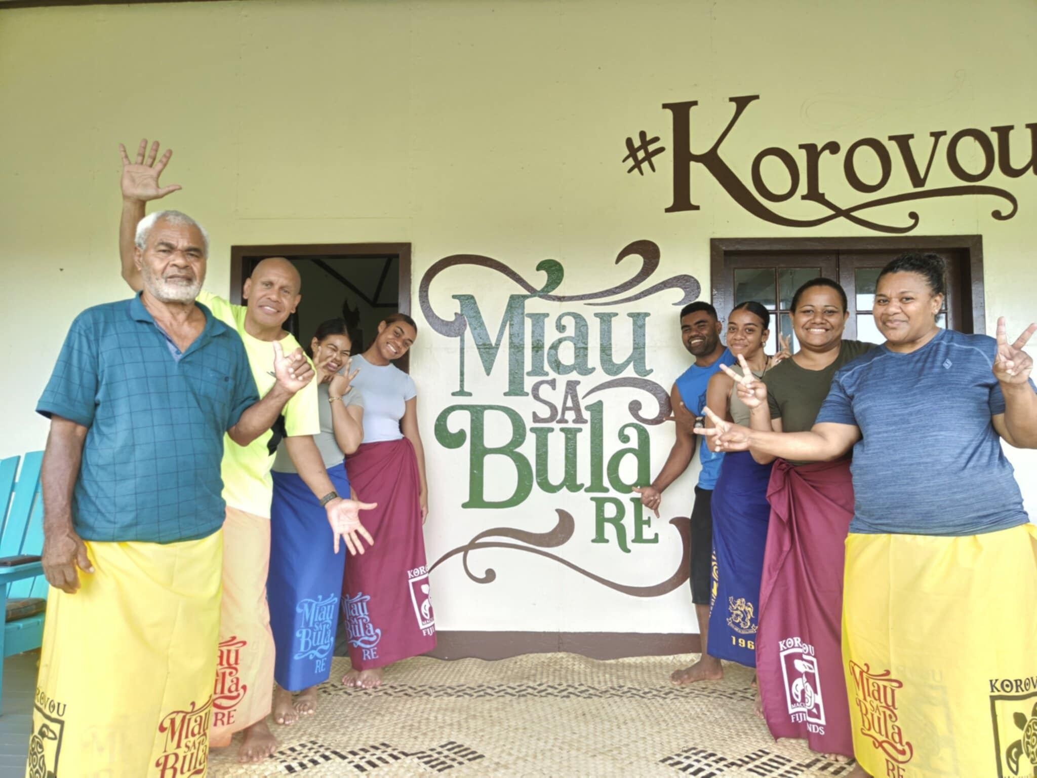 A group of people standing in front of a wall with the text "Miau sa Bulau RE" and "#Korovou".