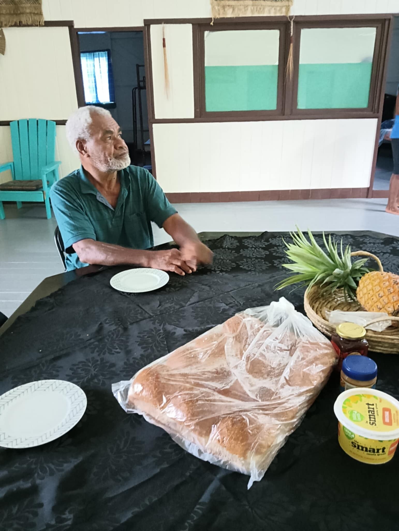 A man seated at a table with a black tablecloth, a plate, a loaf of bread, a pineapple, a basket, and a container of yogurt.