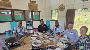 A group of people seated around a large table with various dishes and drinks, in a room with wooden walls and a view of greenery outside. - Korovou Homestay Fiji (Naduri)