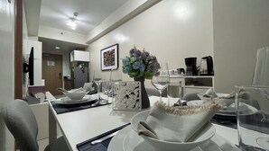 Un espace repas moderne avec une table blanche dressée pour deux personnes, un vase rempli de fleurs et une machine à café en arrière-plan.