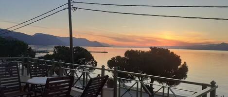 Een terras met tafel en stoelen met uitzicht op een rustig waterlichaam en verre bergen tijdens zonsondergang.