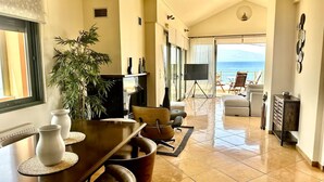 Un vaste espace de vie comprenant une table à manger, des chaises, une grande plante en pot et une vue sur l’océan, accessible par une porte-fenêtre coulissante.