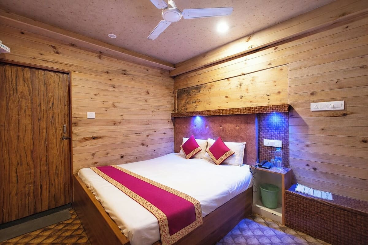 Una habitación con paneles de madera, una cama, un ventilador de techo, una mesita de noche y un cubo de basura.