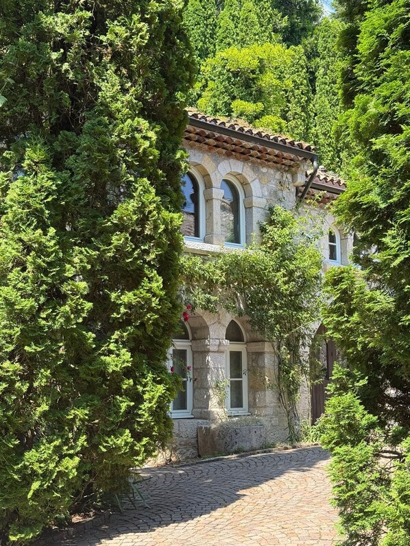 A stone building with arched windows and a tiled roof, surrounded by lush greenery. - Cozy Chalet w/Fireplace, Patio & Barbecue. Pet Friendly! (Gex)
