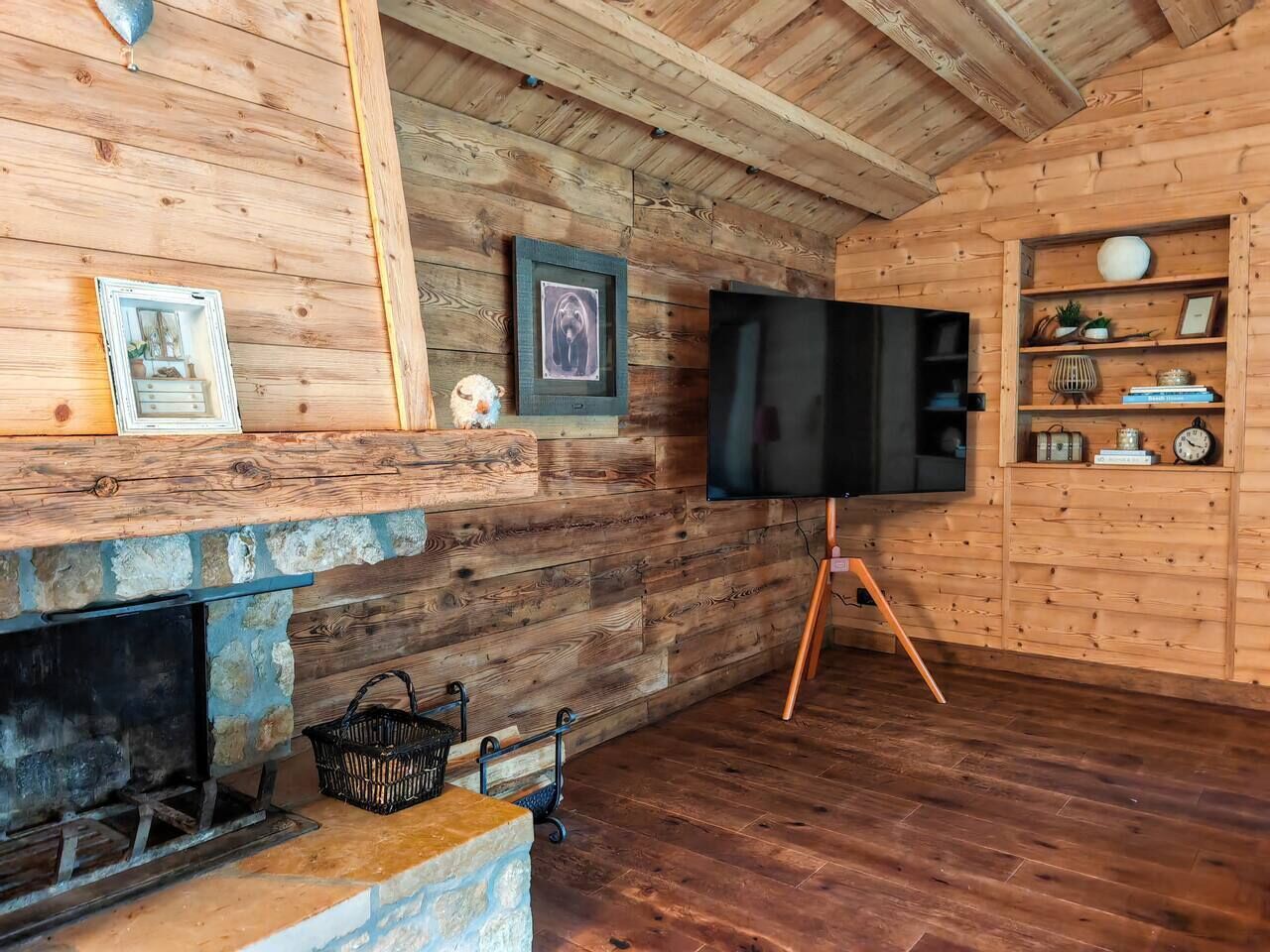 L’intérieur d’un chalet rustique, avec une cheminée en pierre, des murs en bois, un téléviseur posé sur un trépied et une étagère ornée d’objets décoratifs.