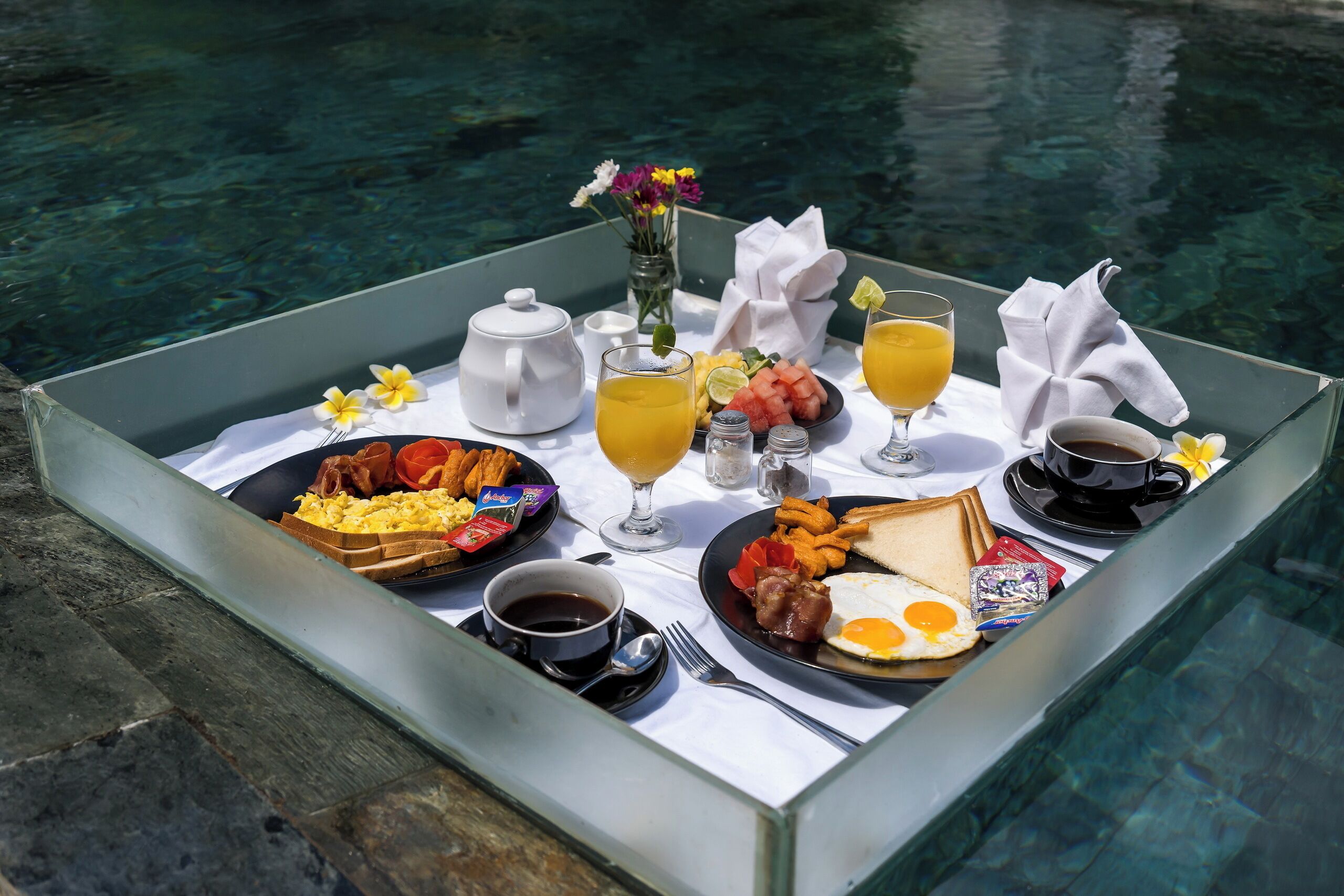A breakfast tray with eggs, bacon, toast, and juice by a pool.