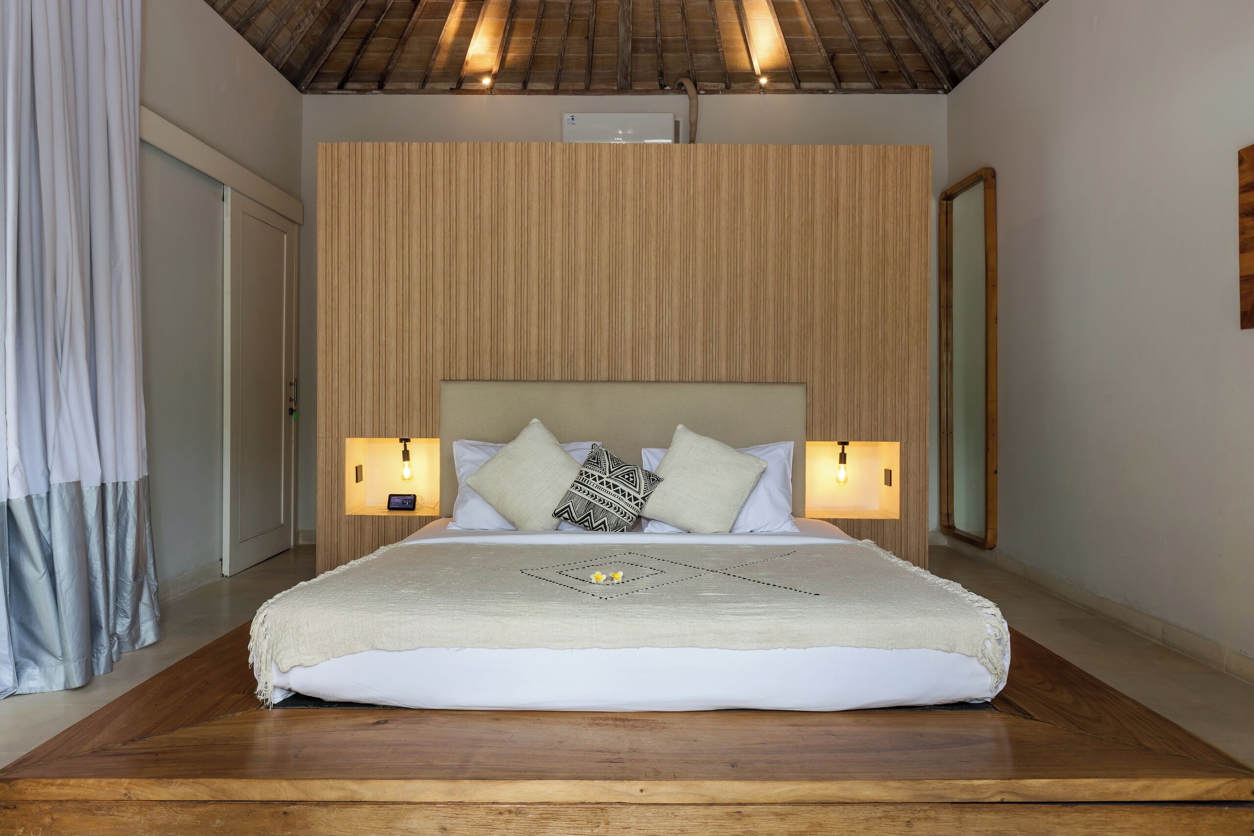 Un dormitorio con cabecera de madera, cama con ropa blanca, dos lámparas de noche y piso de madera.