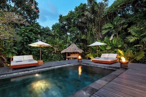 A poolside area with a wooden deck, two lounge chairs, a thatched-roof structure, and a small fire feature. - Villa Reina by Soobali (Kerobokan)
