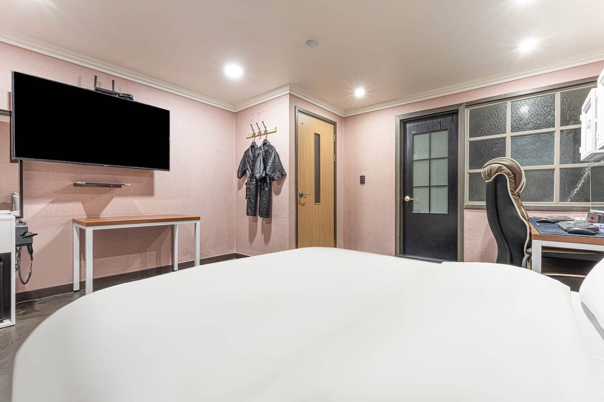 A hotel room with a large bed, a desk with a chair, a television mounted on the wall, and a door with a window.