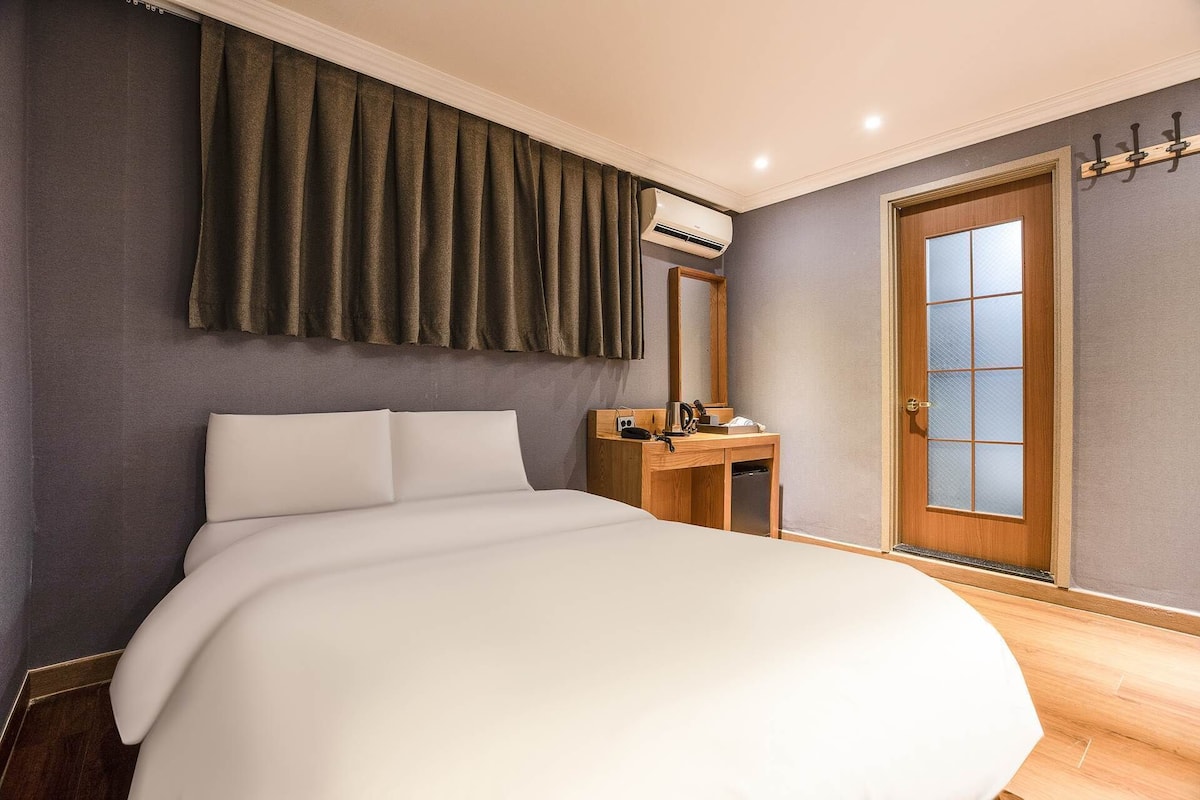 A neatly made bed in a hotel room with a wooden desk, a mirror, and a door with a frosted glass panel.