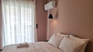 A neatly made bed with white linens and pillows, a window with sheer curtains, and a wall-mounted air conditioner. - Athens Heart Acropolis Balcony (Athens)