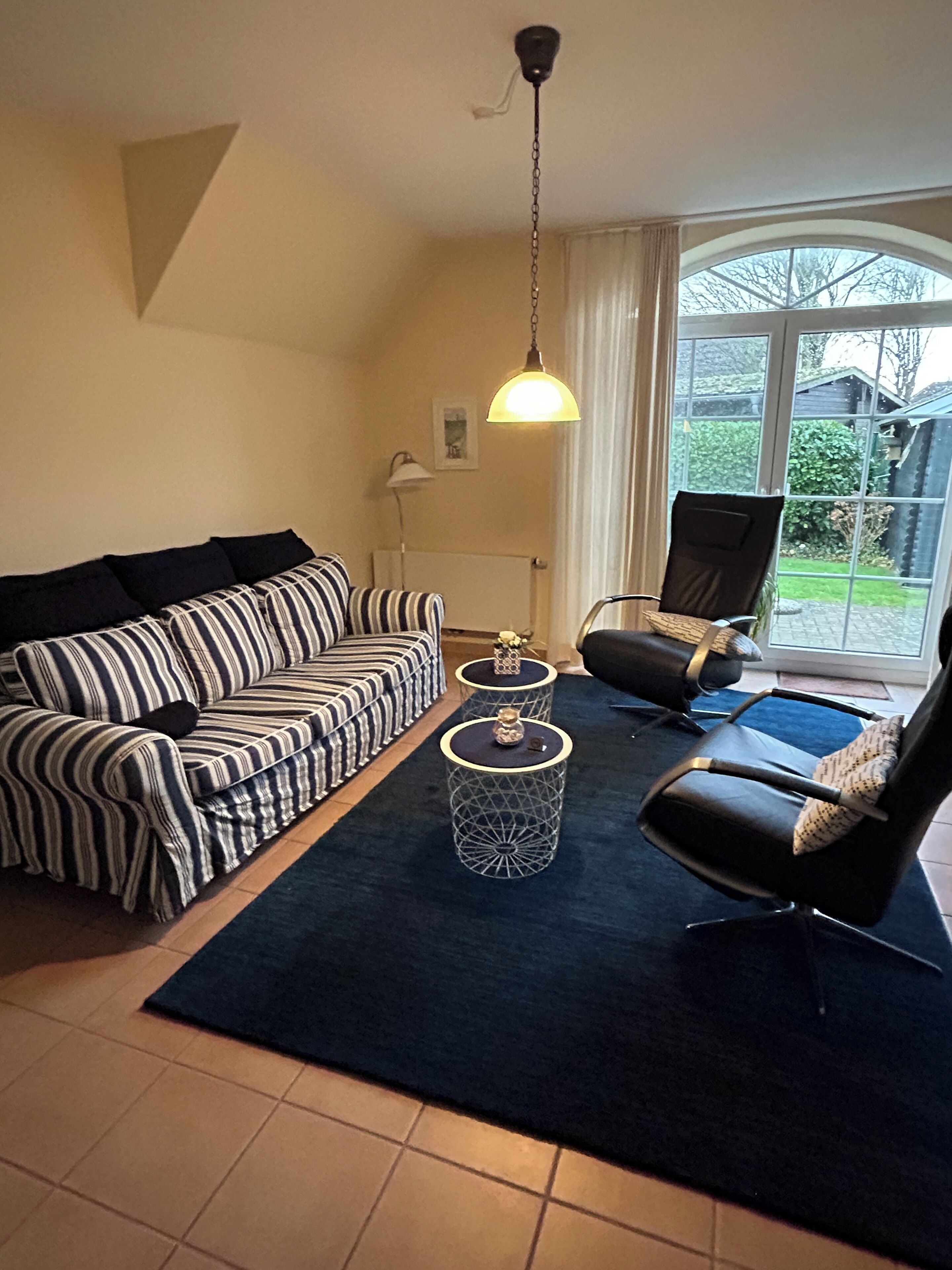 A living room with a striped sofa, a black leather chair, a round coffee table, and a large window with a view of a garden.